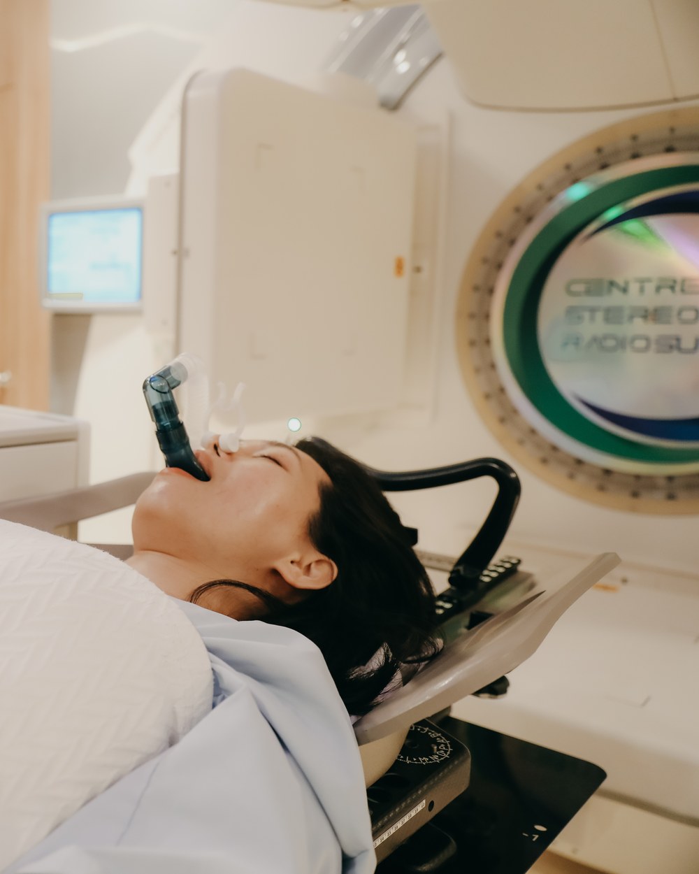 A patient using a breath-hold spirometer device beneath the linear accelerator — the deep-inspiration breath-hold technique used to protect the heart during left-sided breast radiation.